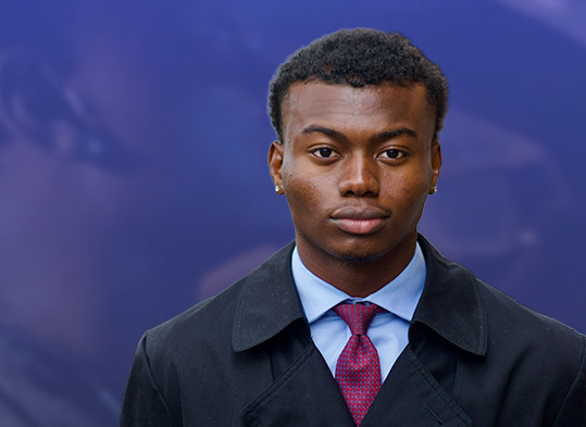 Portrait of a young man in a black coat and red tie, against a blurred blue background. Confident expression.